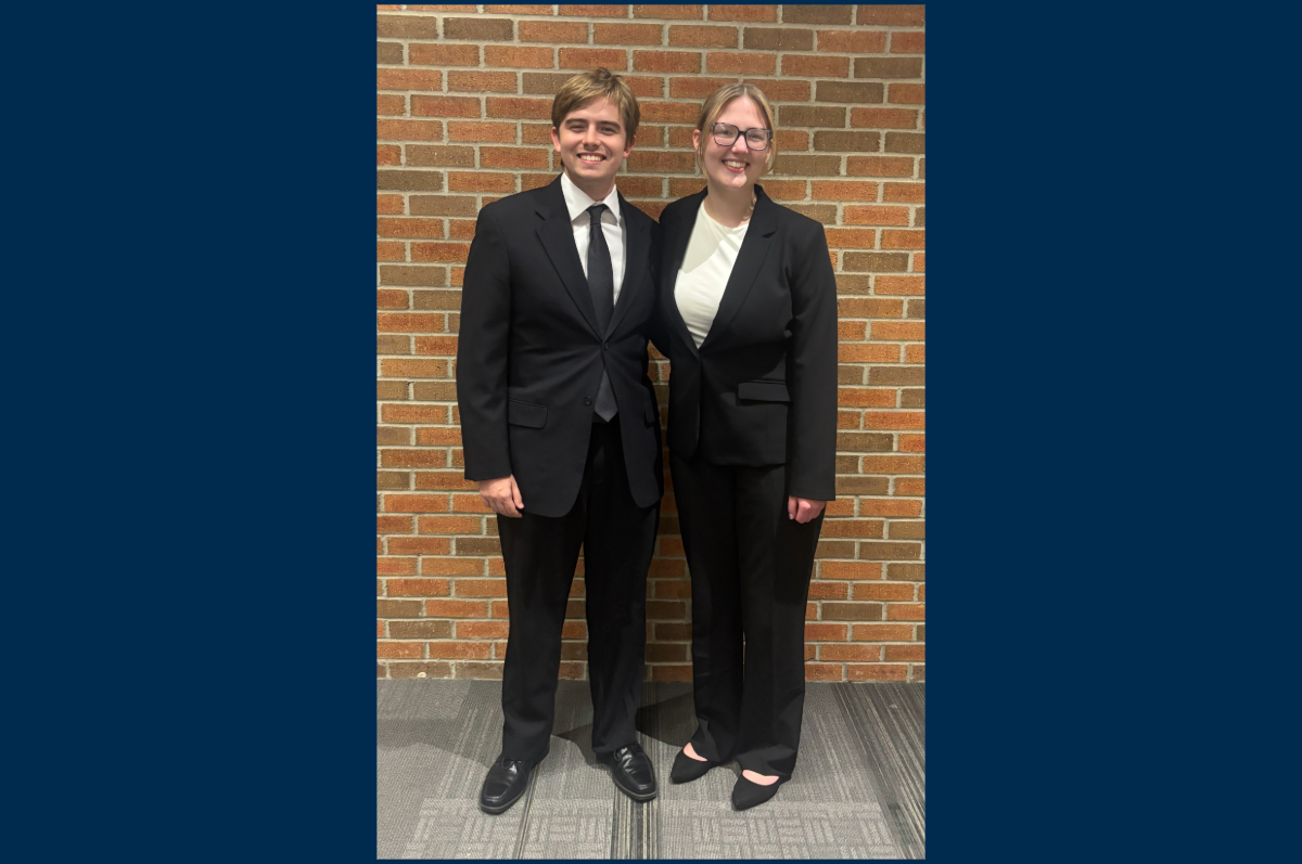 two young adults in black suits stand in front of brick wall